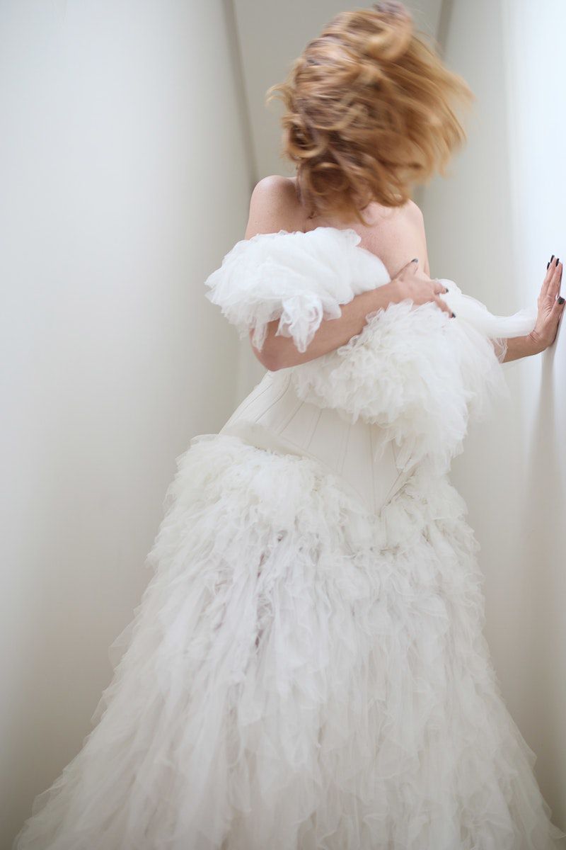 Fashion shot of a woman in a white ruffled dress with blonde hair blurred in motion against a white studio wall in a portrait orientation.