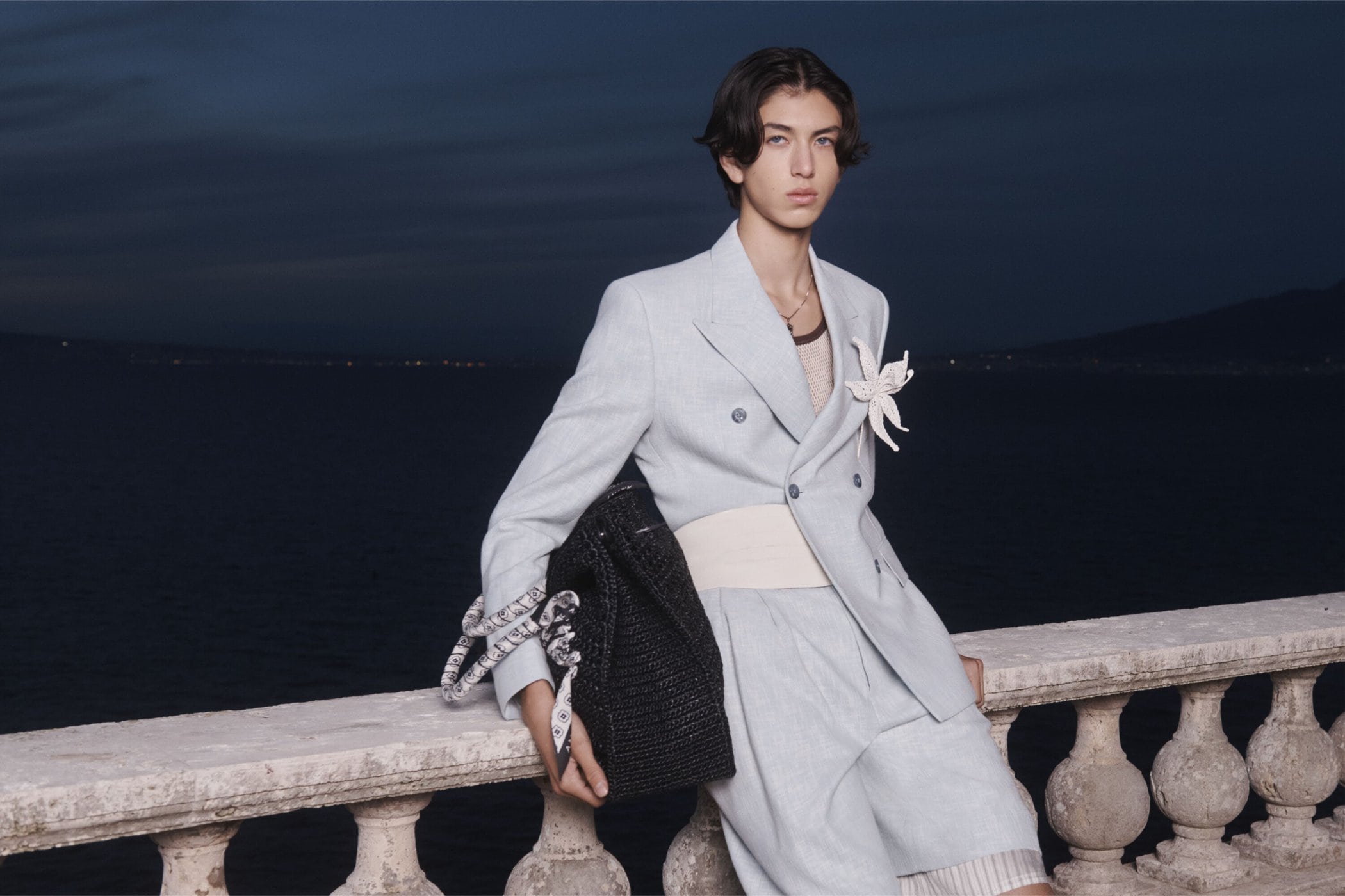 Adult male model wearing a light blue linen suit with a white flower pinned on the jacket poses with a black tote bag outdoors.