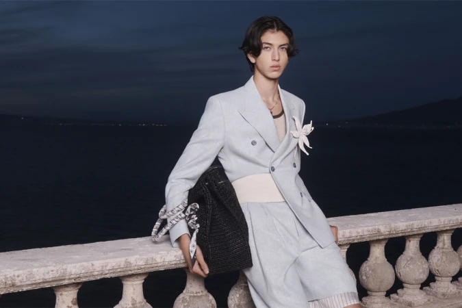 Adult male model wearing a light blue linen suit with a white flower pinned on the jacket poses with a black tote bag outdoors.