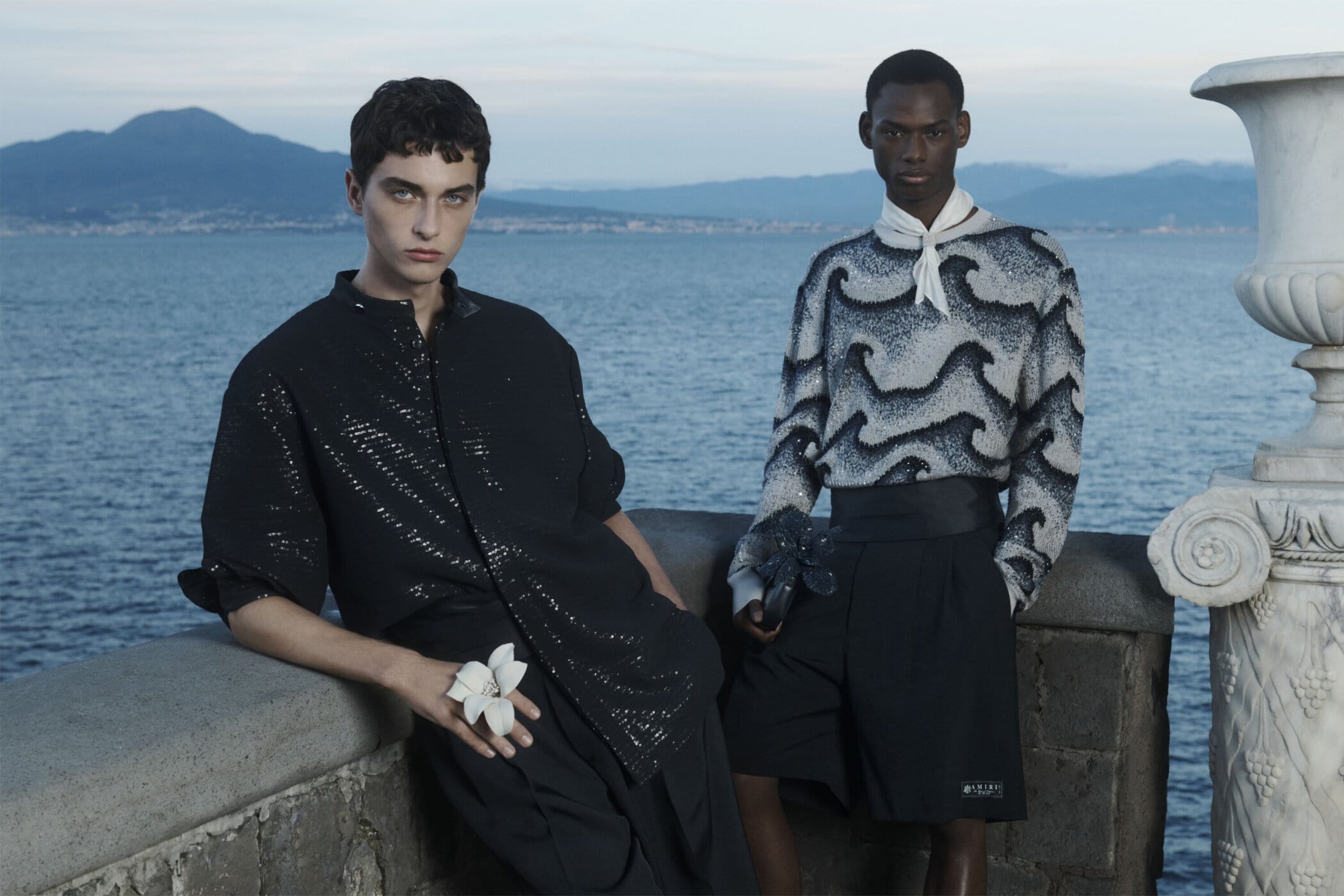 Two fashion models pose on a ledge overlooking the sea, one in a black sequined shirt and the other in a gray wave-patterned sweater.