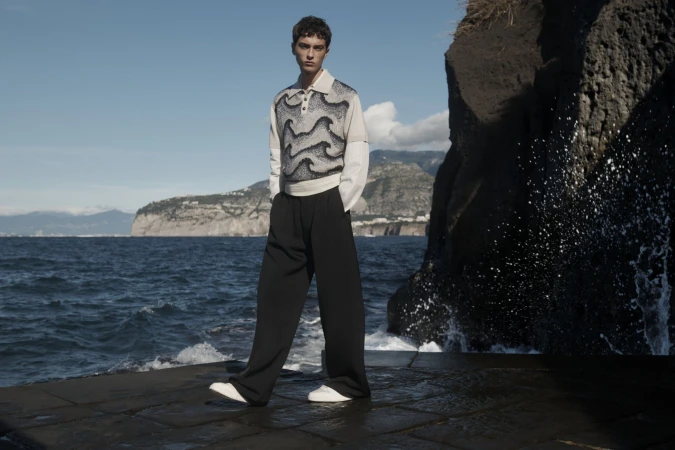 A young man poses in a patterned sweater and wide-leg pants by the sea on a stone pier with a rocky cliff and blue sky.