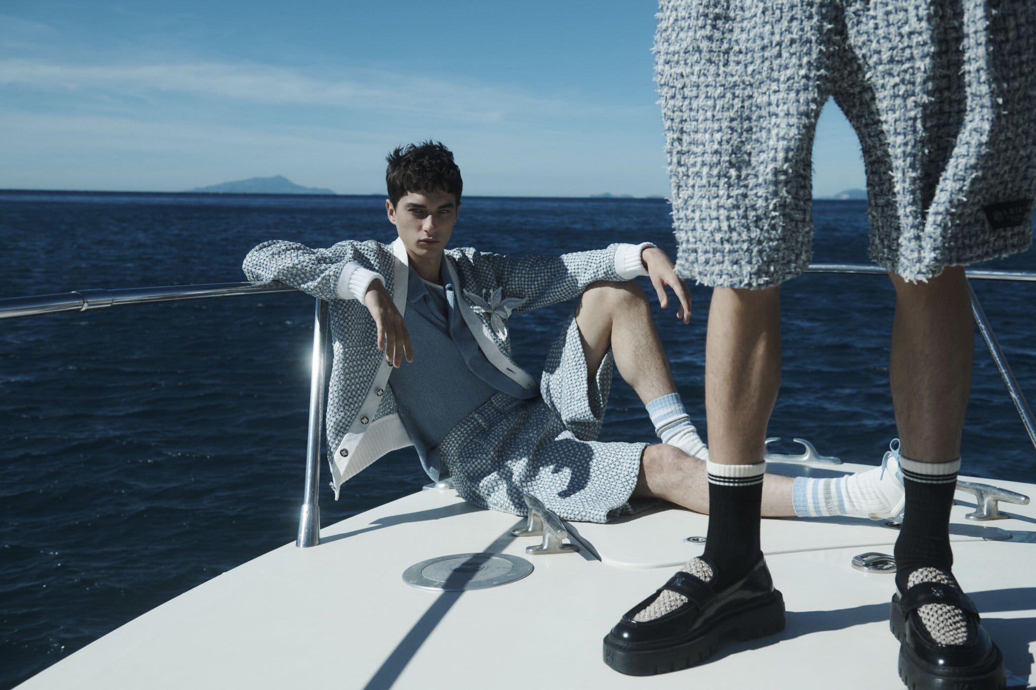 Two men pose on a boat wearing light blue matching outfits in front of a blue ocean and sky, creating a summery fashion scene.