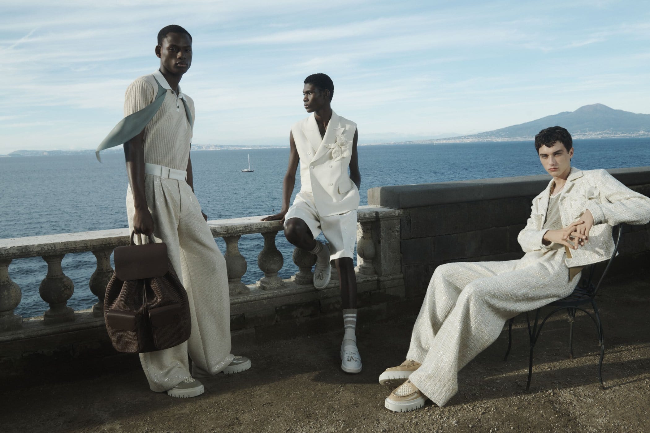 Three adult male fashion models pose outdoors on a terrace overlooking the ocean and mountains, showcasing elegant cream-colored apparel.