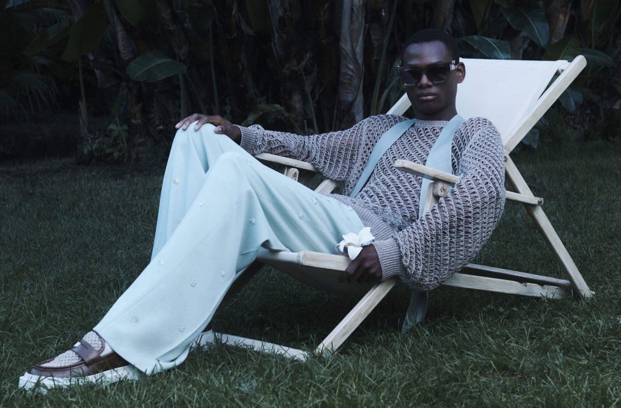 Dark-skinned man in sunglasses sits in a beach chair on grass, wearing knit sweater, light pants with flower details, and brown loafers.