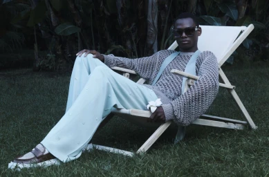 Black man wearing sunglasses and a textured grey knit sweater seated in a white canvas deck chair on lawn grass.
