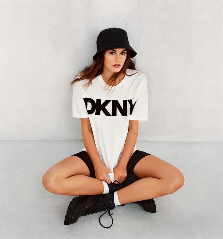 A young adult female poses indoors wearing a DKNY t-shirt, black cycling shorts, boots, and a black bucket hat against a neutral background.