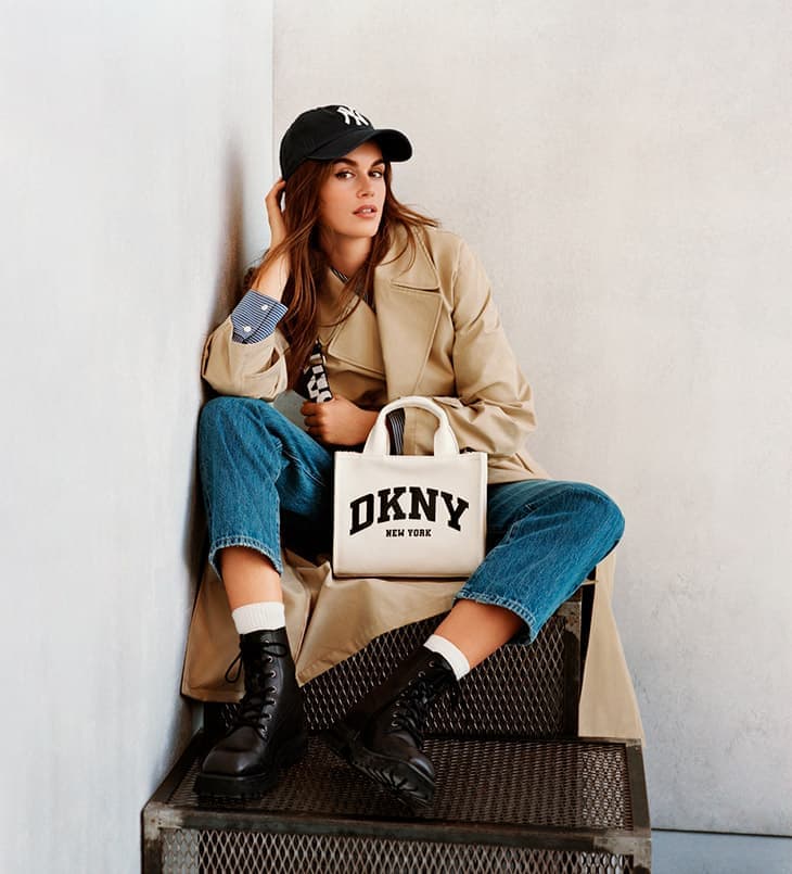 Adult female seated wearing trench coat, baseball cap, jeans, and boots, holding DKNY tote bag indoors against neutral wall.