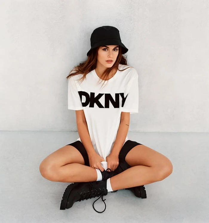 Kaia Gerber wearing a white DKNY t-shirt, black bucket hat, and black boots while seated against a grey backdrop