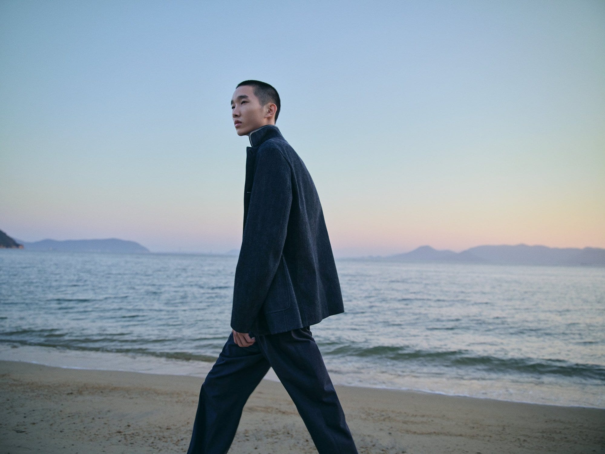 Man walks on beach wearing dark blazer and pants against a soft sunset backdrop with ocean water and hazy coastal mountains.
