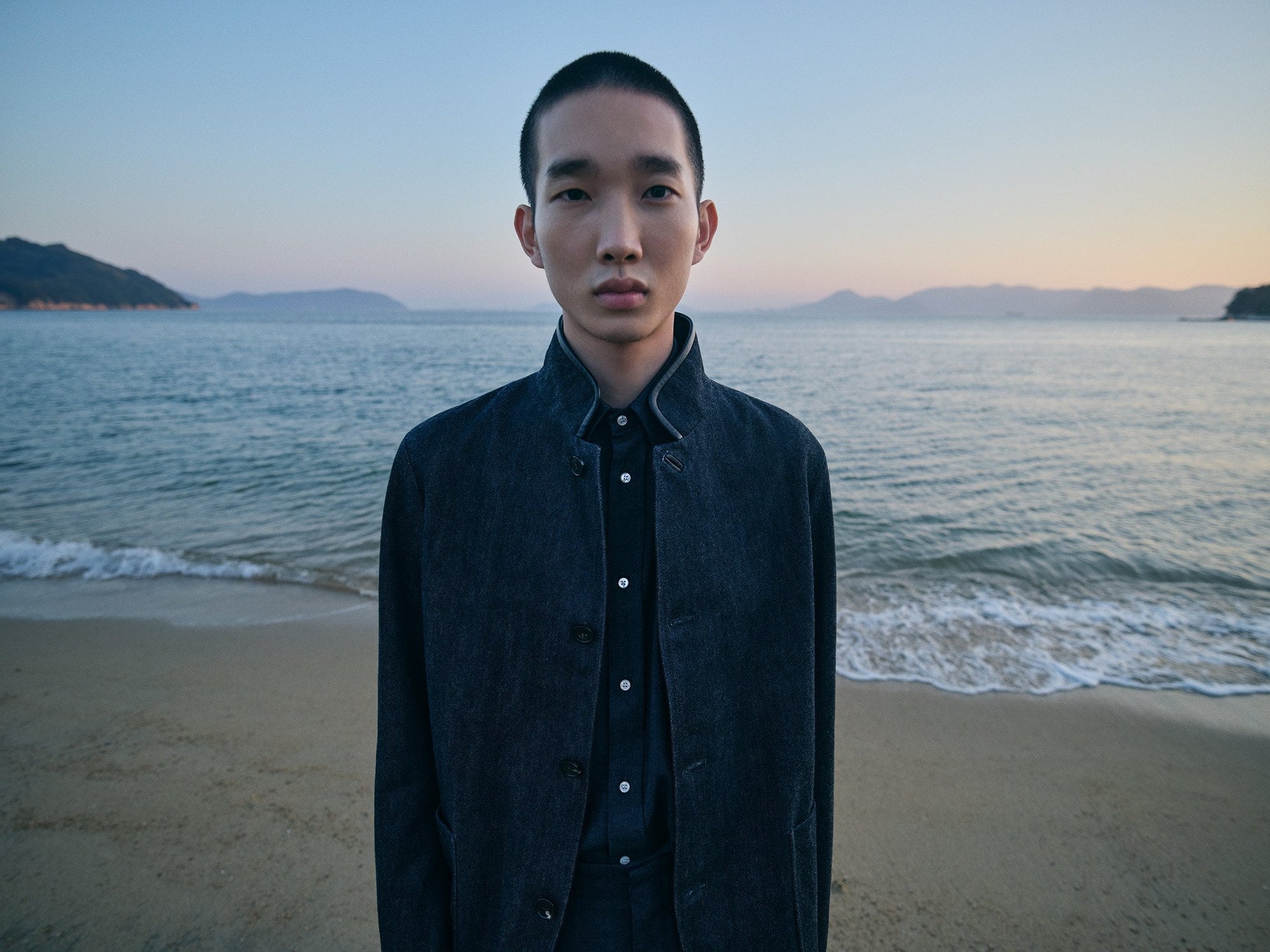 An Asian male model poses on a beach in a dark jacket and shirt, looking directly at the camera with a neutral expression during dusk.