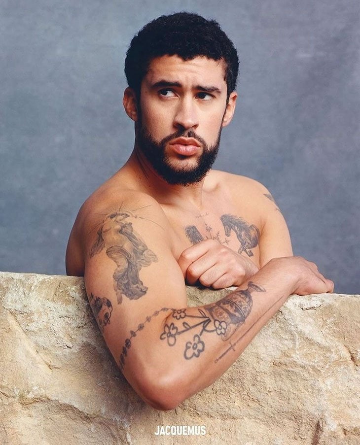 Portrait of a man with dark hair and tattoos, posing shirtless in front of a stone wall with a gray background, designer logo visible.