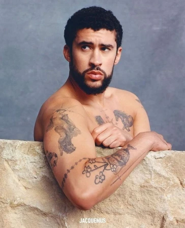 Portrait of a man with dark hair and tattoos, posing shirtless in front of a stone wall with a gray background, designer logo visible.