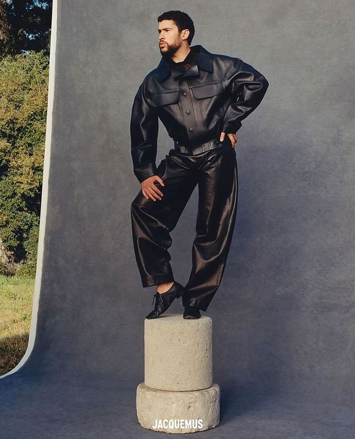 Adult male posing in a black leather jacket and pants, standing on stacked stone pedestals in an indoor photo studio with neutral backdrop.