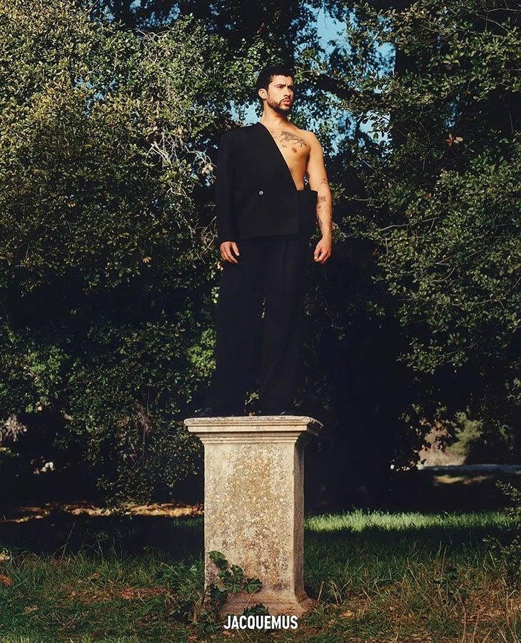 Man standing on a plinth with jacket draped over one shoulder in a verdant outdoor setting, wearing dark trousers. Jacquemus logo visible.
