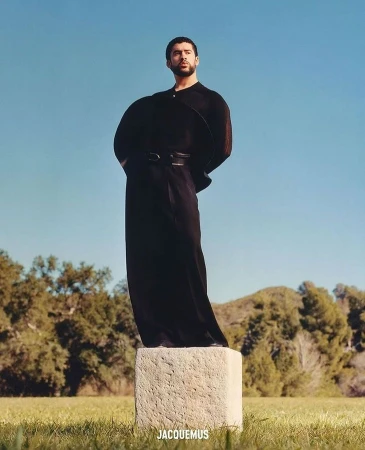 An adult man in a black outfit poses on a concrete block in a grassy field under a clear blue sky, with trees and hills in the background.