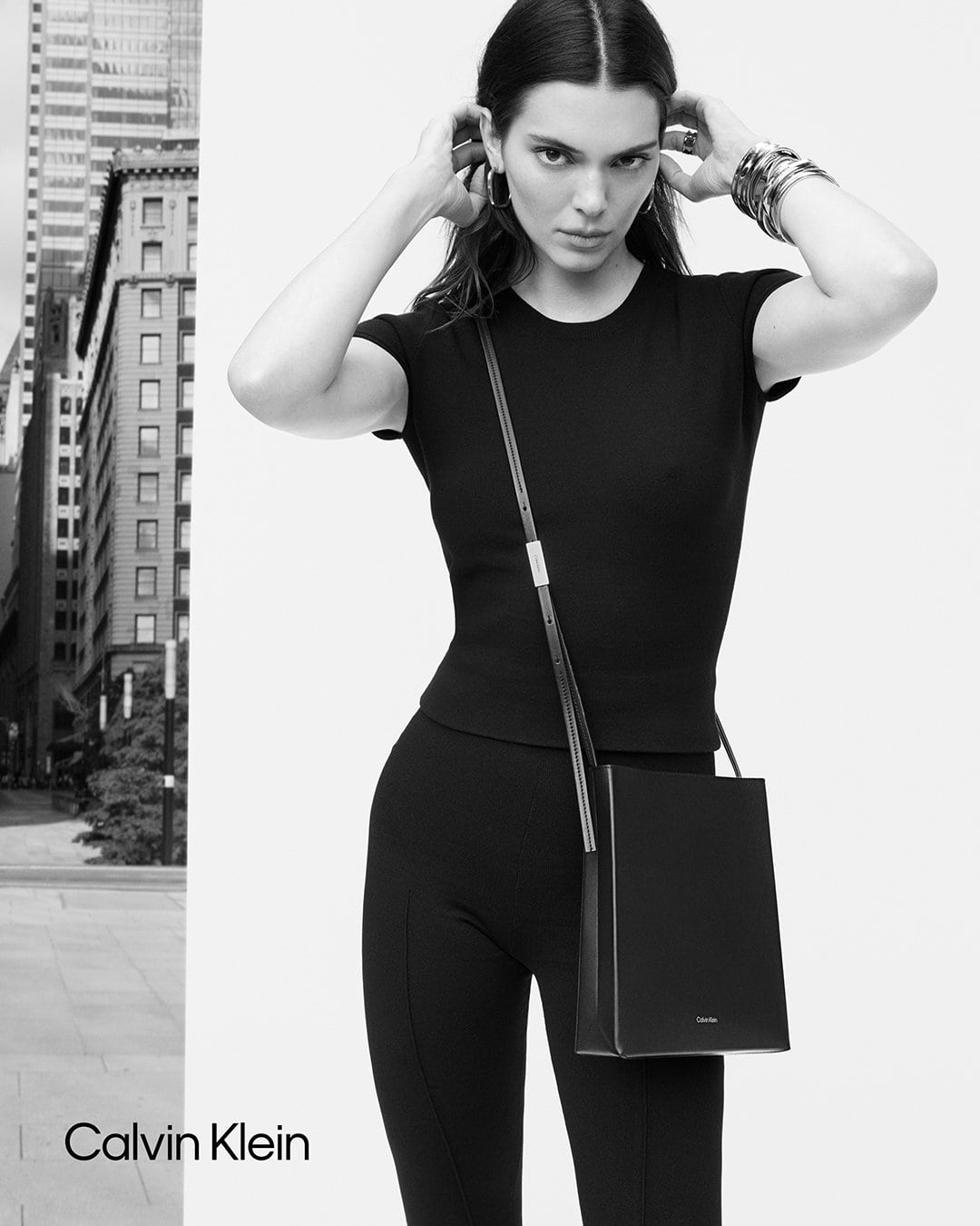 Kendall Jenner poses in a black top and leggings, wearing hoop earrings and bracelets, with a Calvin Klein purse in a city setting.