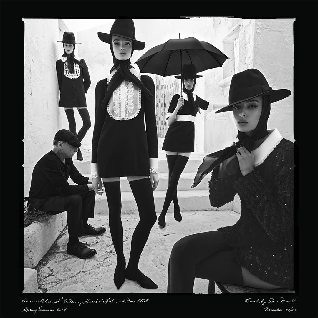 Black-and-white fashion shot featuring five models in vintage-style black dresses and hats, staged in a stepped outdoor setting.