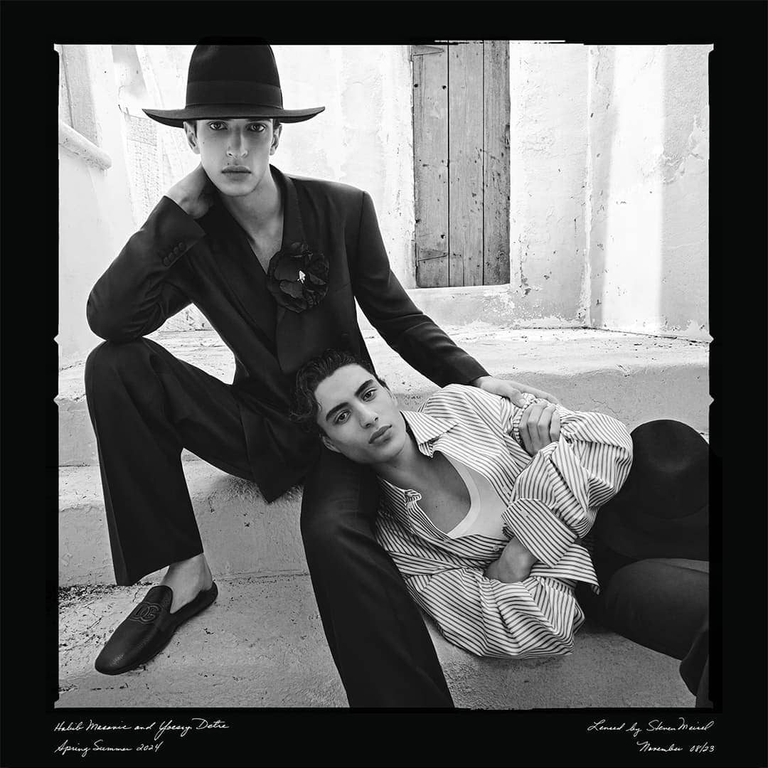 Two men in fashionable suits pose on concrete steps in a black and white editorial fashion shot with a minimalist backdrop and door.
