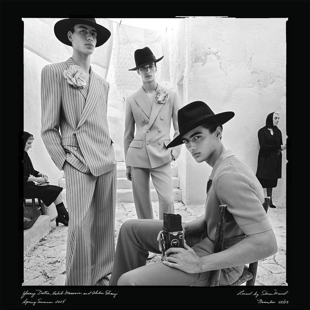 Black and white fashion campaign image featuring three male models wearing hats and tailored suits against a rustic architectural backdrop.