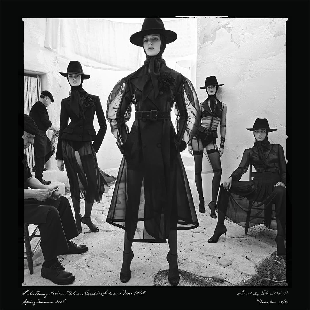 Black and white fashion shot with five models wearing hats, sheer outerwear, and dark clothing in an outdoor setting with stone walls.