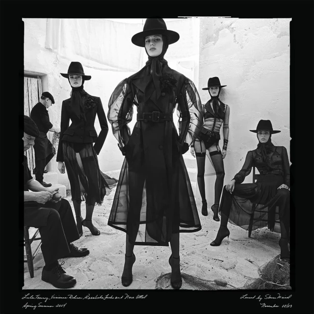 Black and white fashion shot with five models wearing hats, sheer outerwear, and dark clothing in an outdoor setting with stone walls.