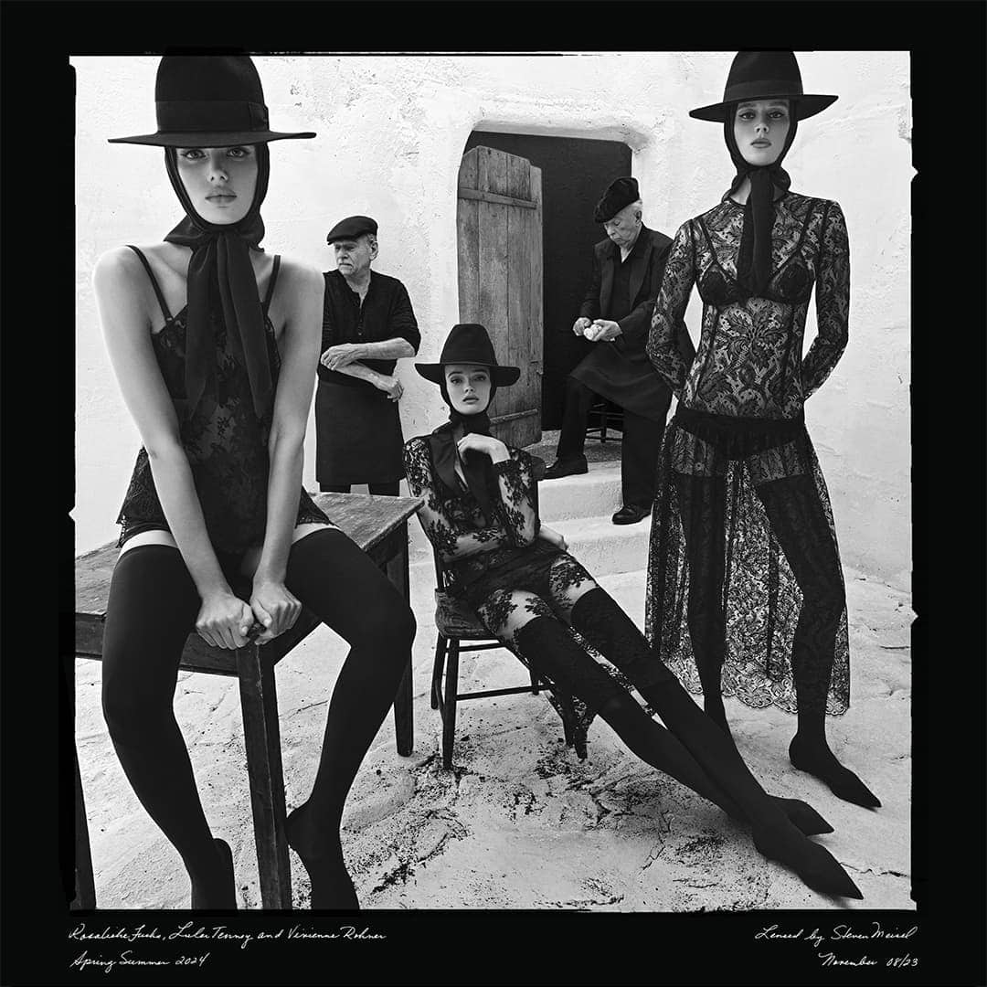 Black and white fashion shot of three women wearing lace outfits and hats with two older men in the background in a courtyard.