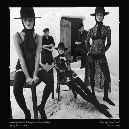 Black and white fashion shot of three women wearing lace outfits and hats with two older men in the background in a courtyard.