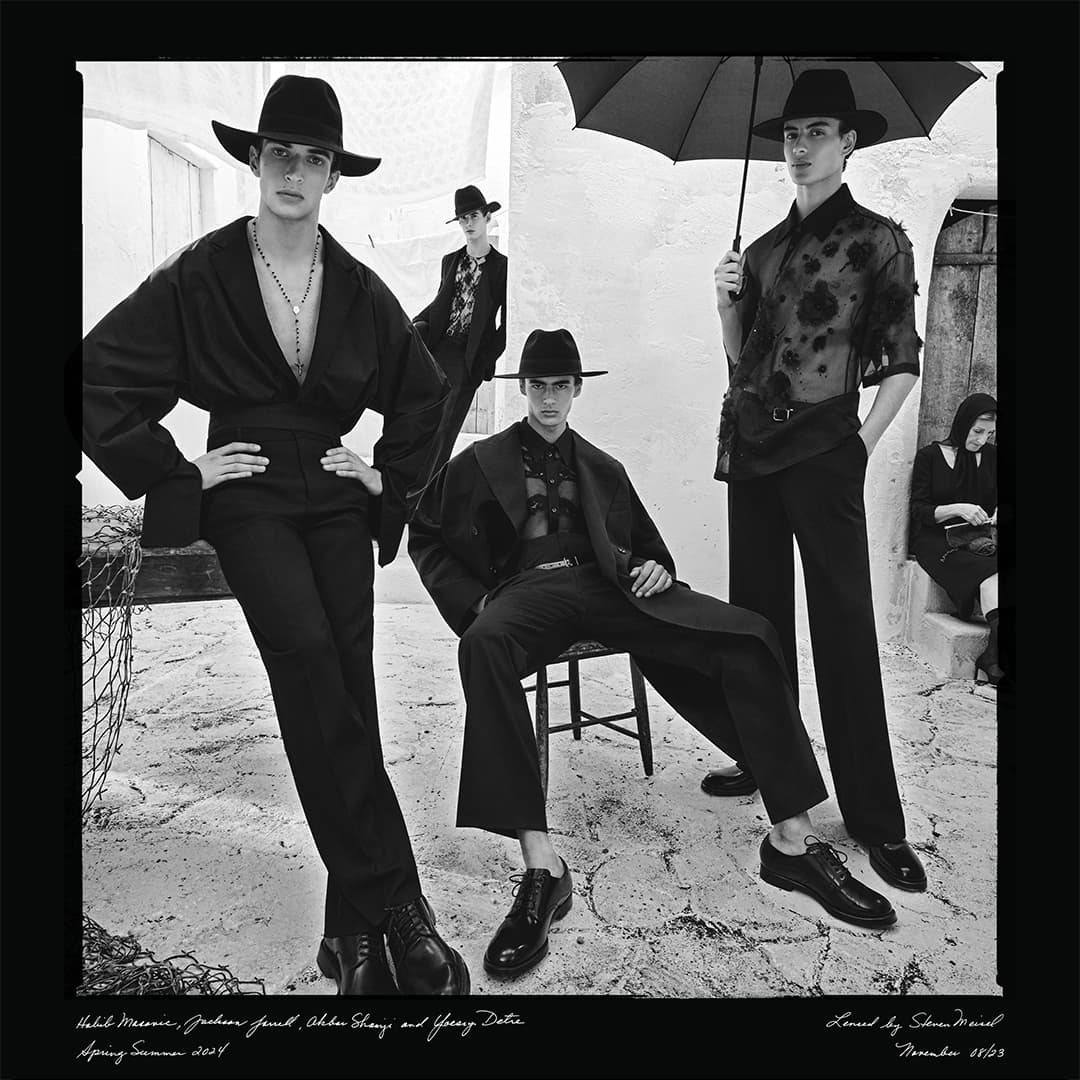 Black and white fashion shot of five adult males wearing hats and dark clothing, posing outside on a stone surface with a building backdrop.