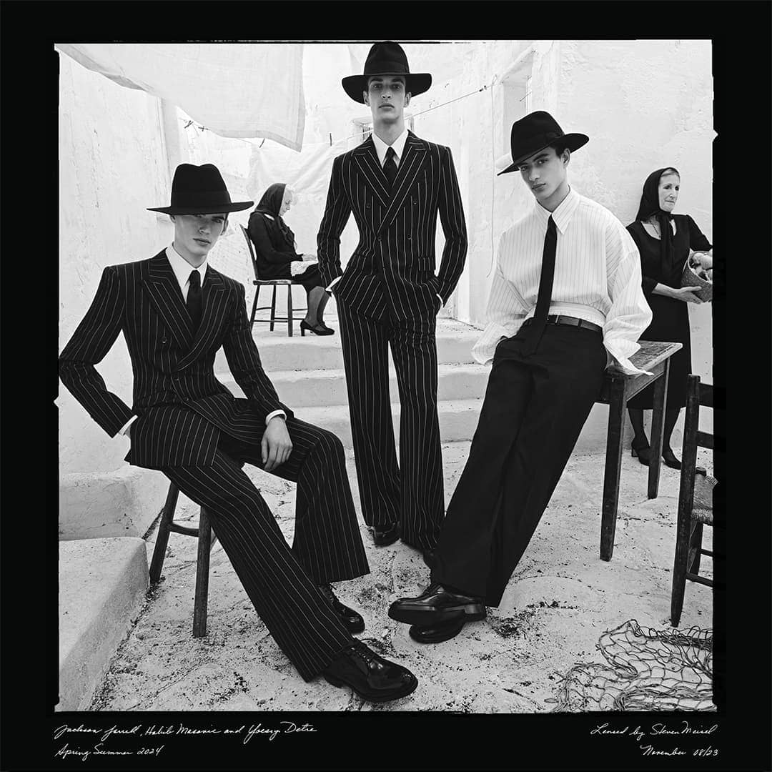 Black and white fashion photography showcasing three young male models in pinstripe suits and hats, with seated women in the background.