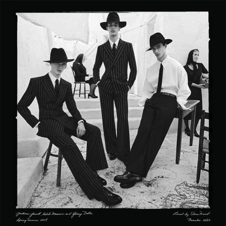 Black and white fashion photography showcasing three young male models in pinstripe suits and hats, with seated women in the background.