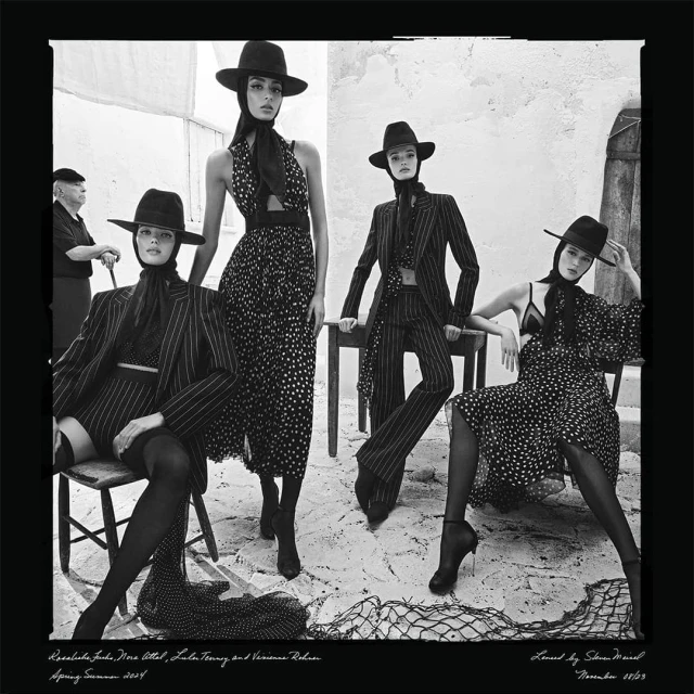 Black and white fashion editorial with four women models in patterned dresses and suits, wearing hats, set against a textured wall.