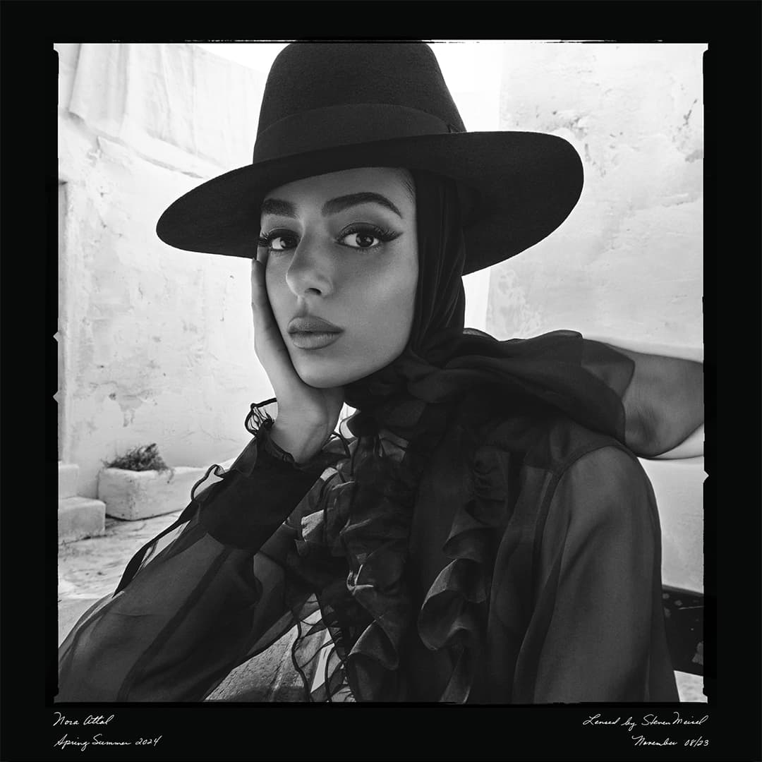 A portrait of a young woman with dark hair and a hat. She rests her face on her hand, and poses in front of a white stucco wall.