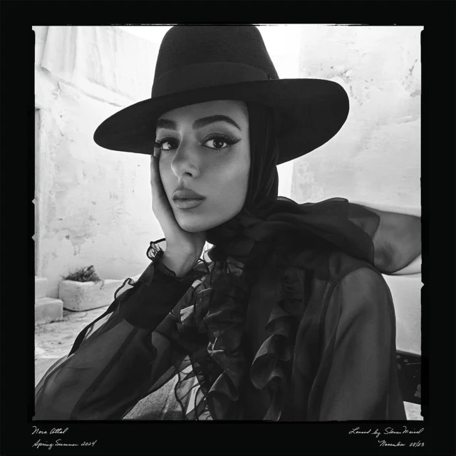 A portrait of a young woman with dark hair and a hat. She rests her face on her hand, and poses in front of a white stucco wall.