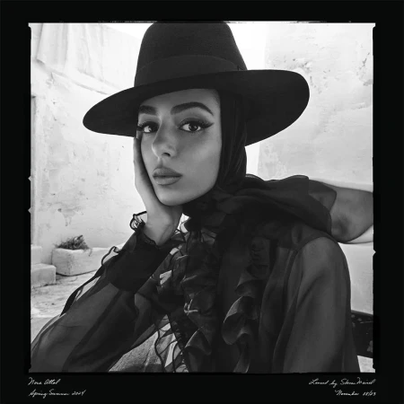 A portrait of a young woman with dark hair and a hat. She rests her face on her hand, and poses in front of a white stucco wall.