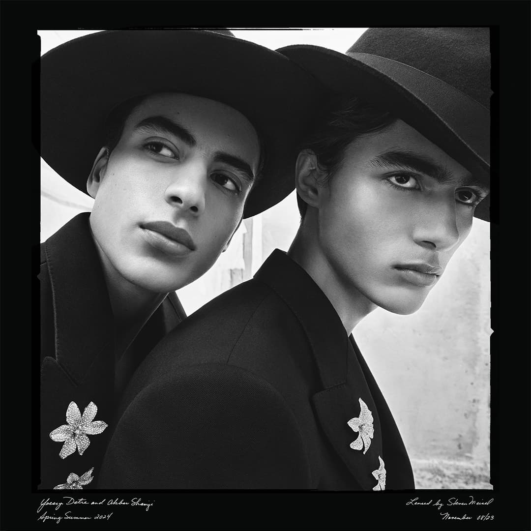Black-and-white portrait of two dark-haired men wearing hats and jackets with floral brooches, set against a blurred background.