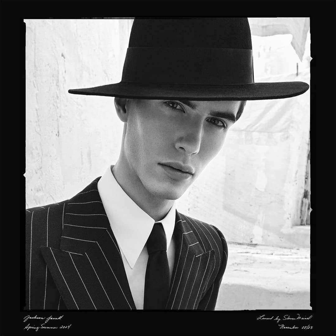 Black and white fashion portrait of a man wearing a pinstripe suit and a black hat, posing against a textured wall, shot at eye-level.