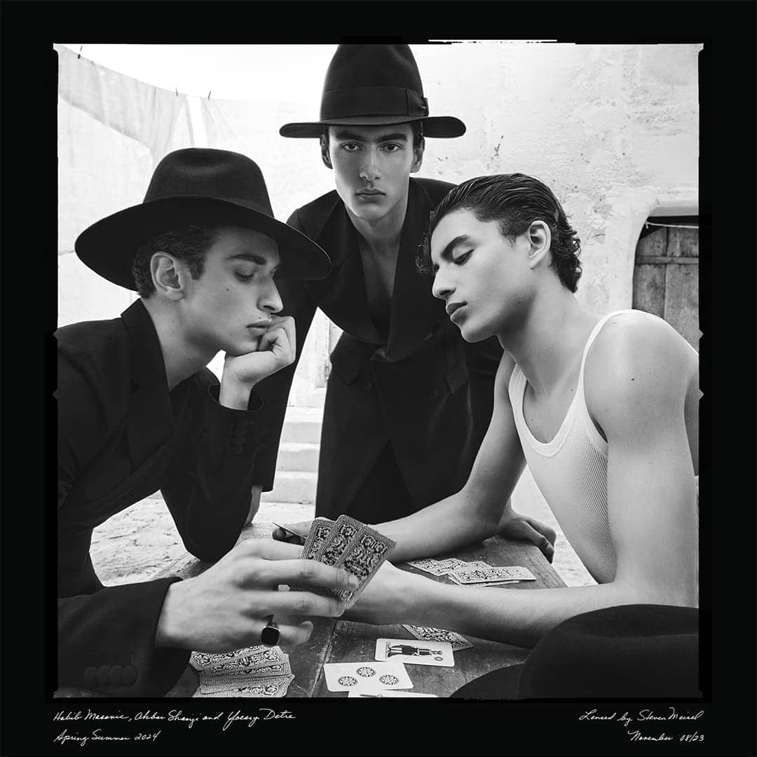 Black and white portrait of three young men wearing hats and dark clothing sitting at a table with playing cards, outdoors, high fashion.