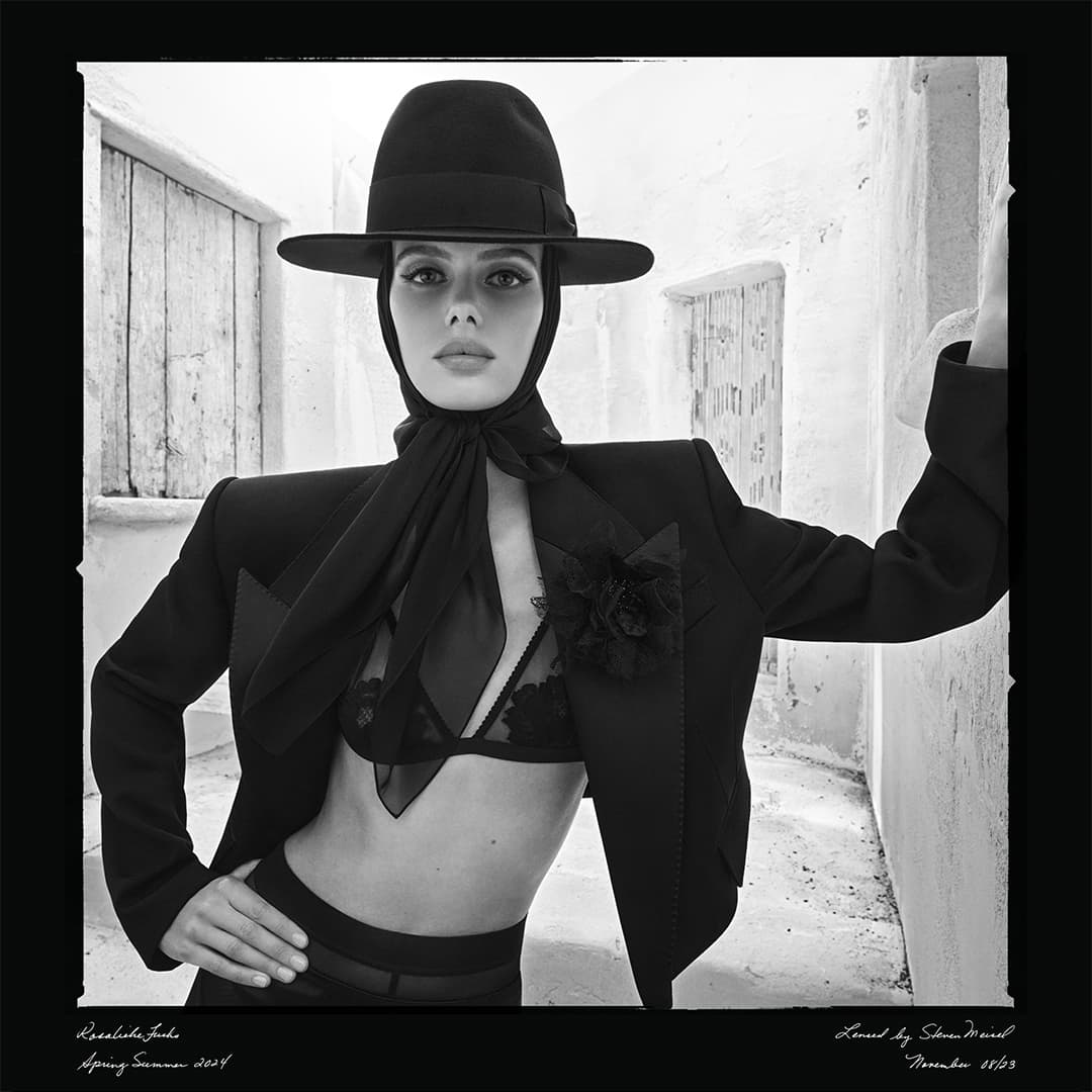 Woman poses in black bra, blazer, and hat against a white stone wall, showcasing a high-fashion editorial style in monochrome.