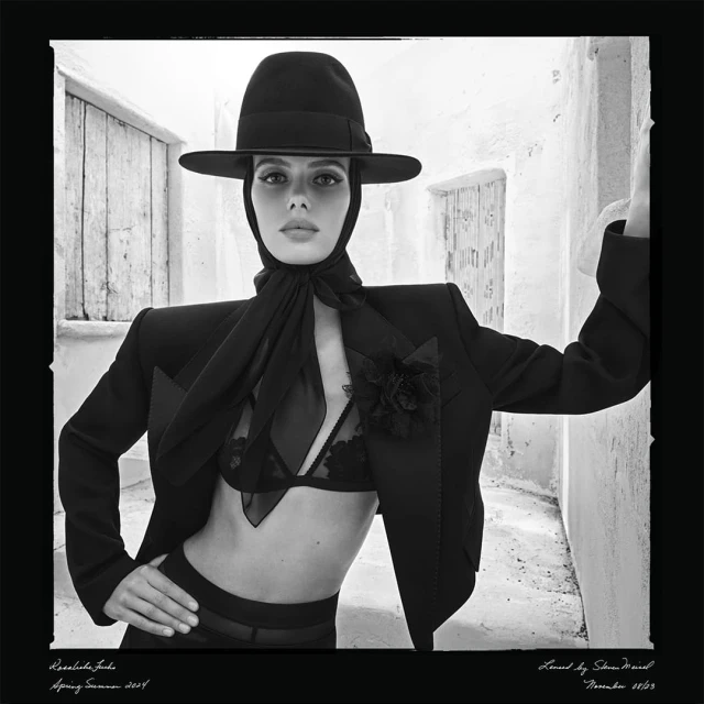 Woman poses in black bra, blazer, and hat against a white stone wall, showcasing a high-fashion editorial style in monochrome.