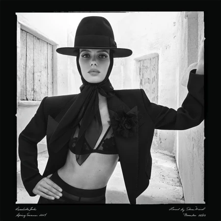 Woman poses in black bra, blazer, and hat against a white stone wall, showcasing a high-fashion editorial style in monochrome.