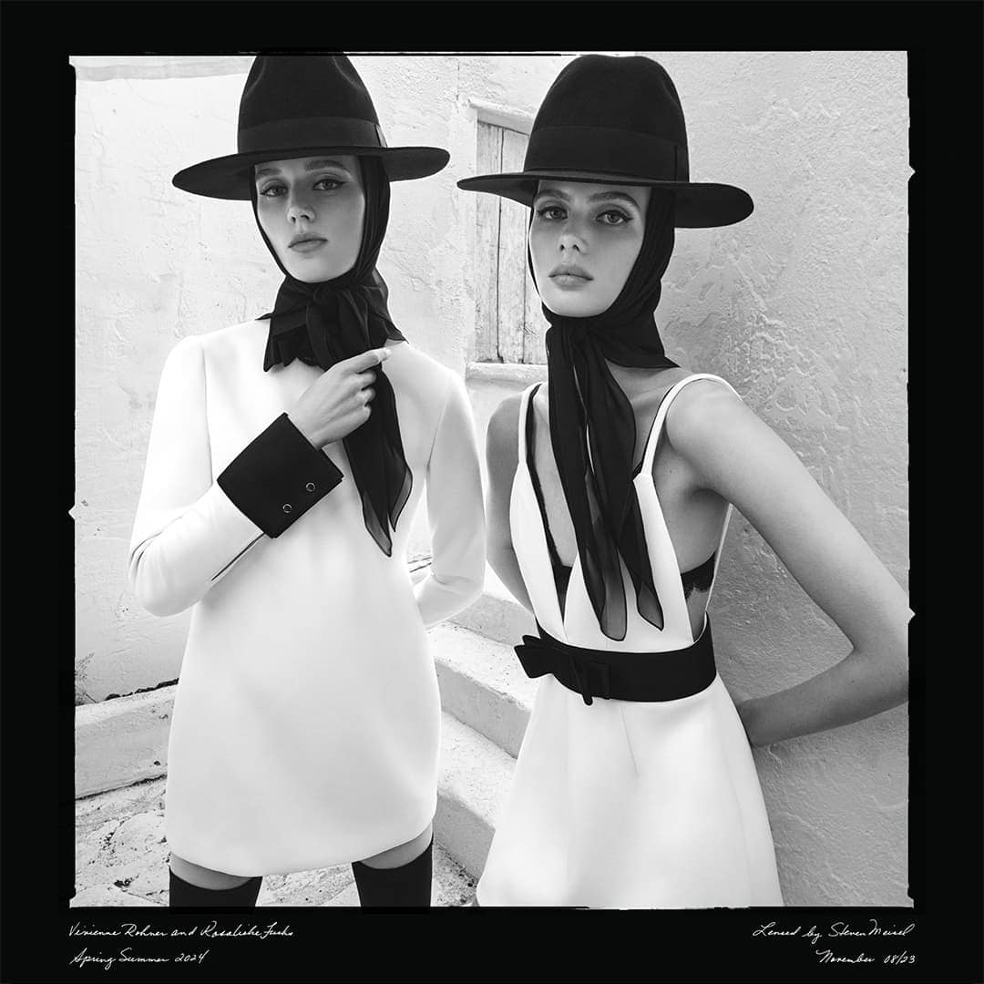 Two women in white dresses and black hats pose on steps in a black and white fashion shot, showcasing elegant and classic style.