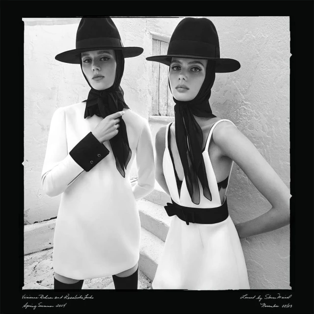 Two women in white dresses and black hats pose on steps in a black and white fashion shot, showcasing elegant and classic style.