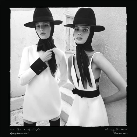 Two women in white dresses and black hats pose on steps in a black and white fashion shot, showcasing elegant and classic style.