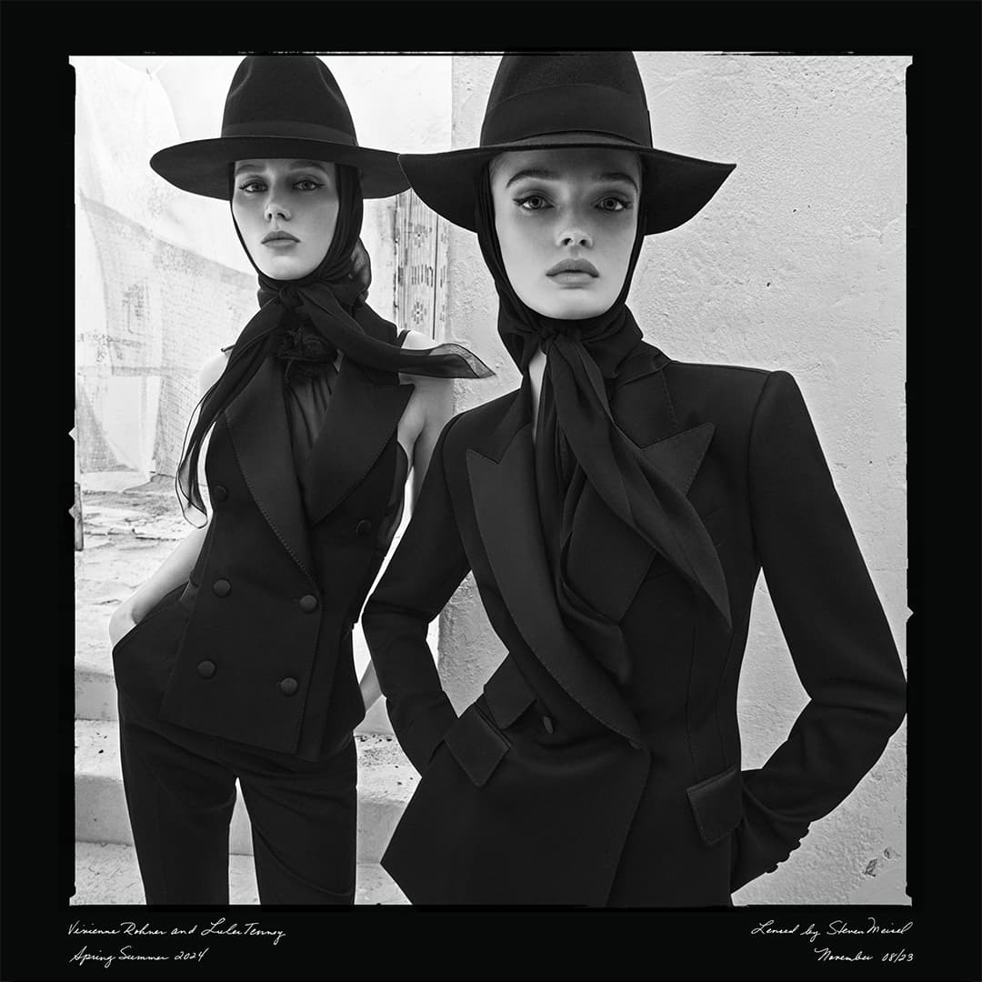 Two women pose in black suits and hats in a black and white fashion editorial shot outdoors against a textured wall backdrop.