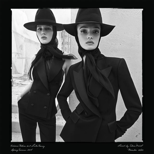 Two women pose in black suits and hats in a black and white fashion editorial shot outdoors against a textured wall backdrop.