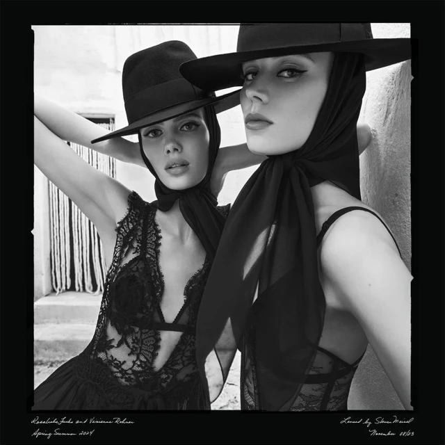 Black and white portrait of two adult women in hats and lace tops posing closely together against a textured wall in outdoor daylight.