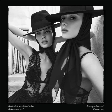 Black and white portrait of two adult women in hats and lace tops posing closely together against a textured wall in outdoor daylight.