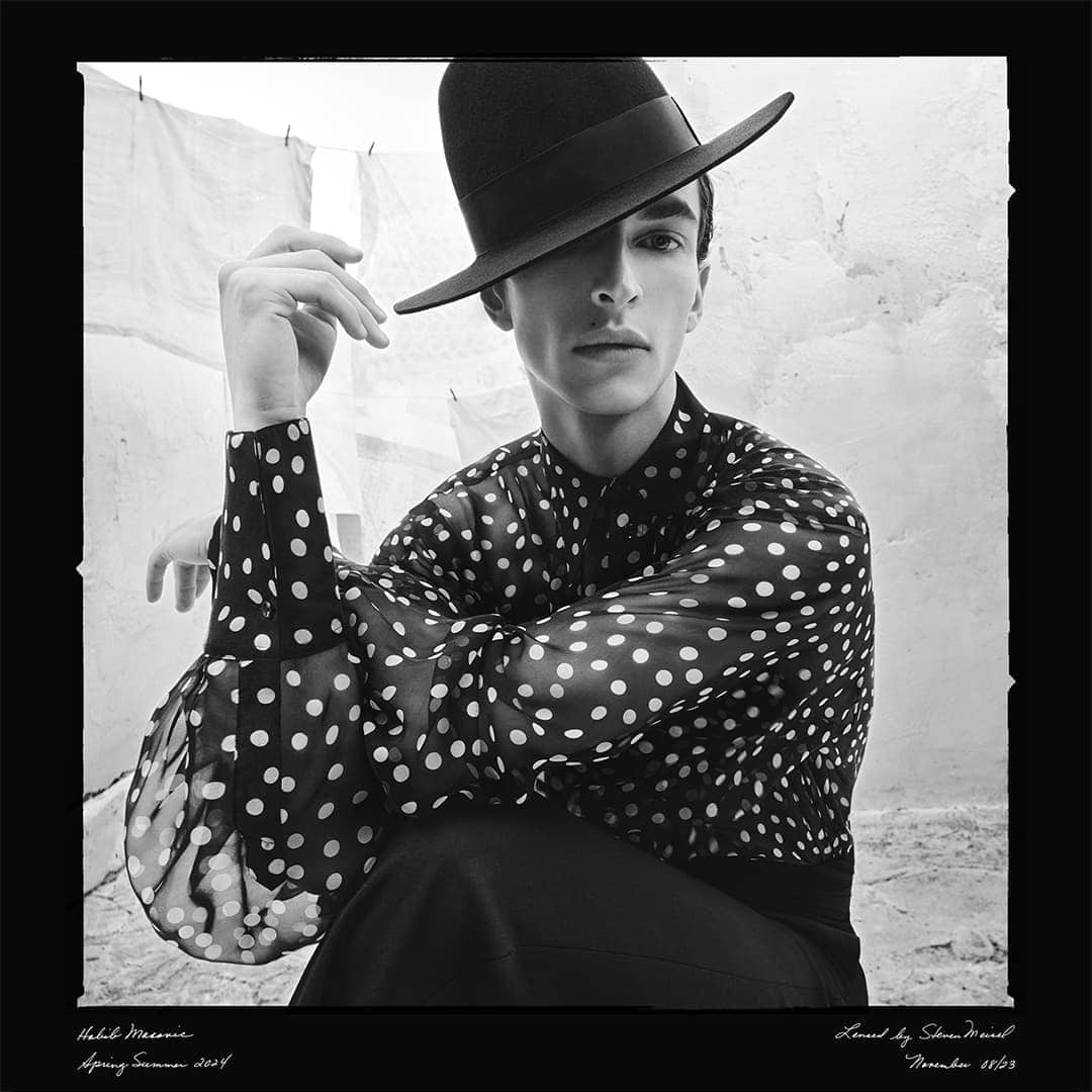 Monochrome fashion portrait of a young adult man in a polka dot shirt and wide-brimmed hat, seated against a textured wall backdrop with laundry line.