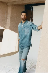 Man wearing a denim shirt and blue jeans leaning against a concrete pillar in front of a white studio backdrop.