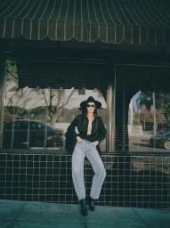 Model wearing blue jeans, black shirt, and hat leaning against a store window in Los Angeles for 7 For All Mankind